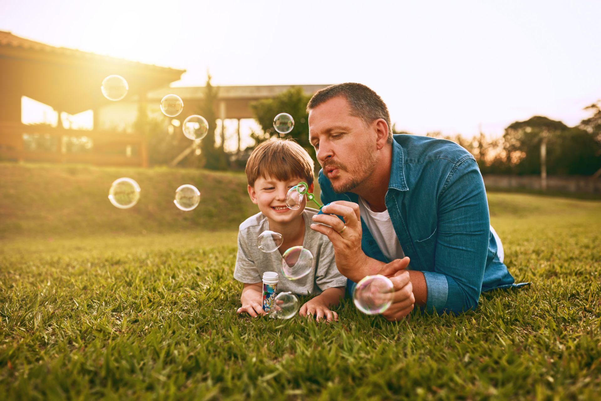 Father and son lying on grass, blowing bubbles joyfully in a sunny outdoor setting.